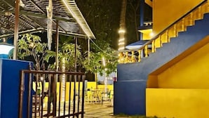 A gated outdoor area with a tiled floor, a blue fence, and a yellow building with a staircase. - Karma Stays Hostel Morjim (Morjim)