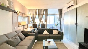 Una sala de estar moderna con un sofá, una mesa de centro y un televisor. Hay una ventana con cortinas, una lámpara y un estante con adornos.