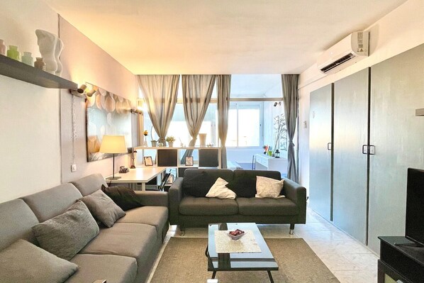 A modern living room with a sofa, a coffee table, and a TV. There is a window with curtains, a lamp, and a shelf with decorative items.