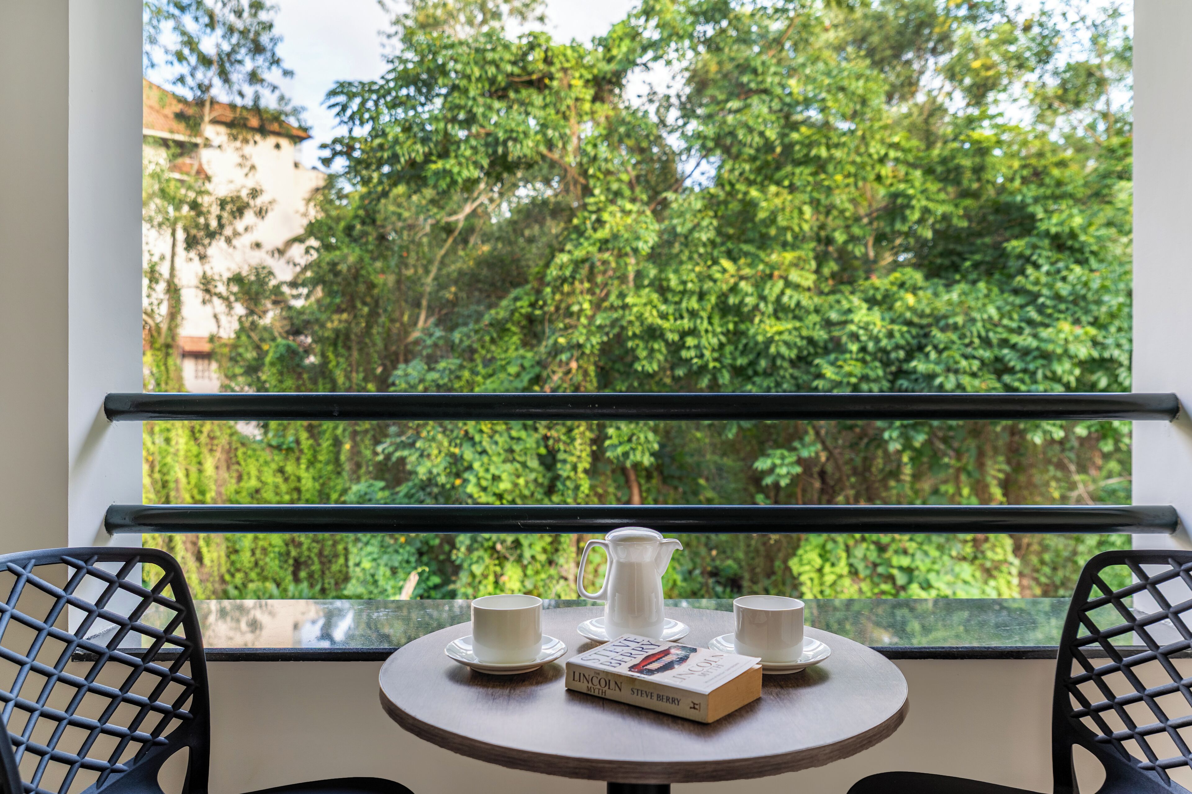 Mesa redonda con un libro, una jarrón y dos tazas en un balcón con vista a follaje verde exuberante.