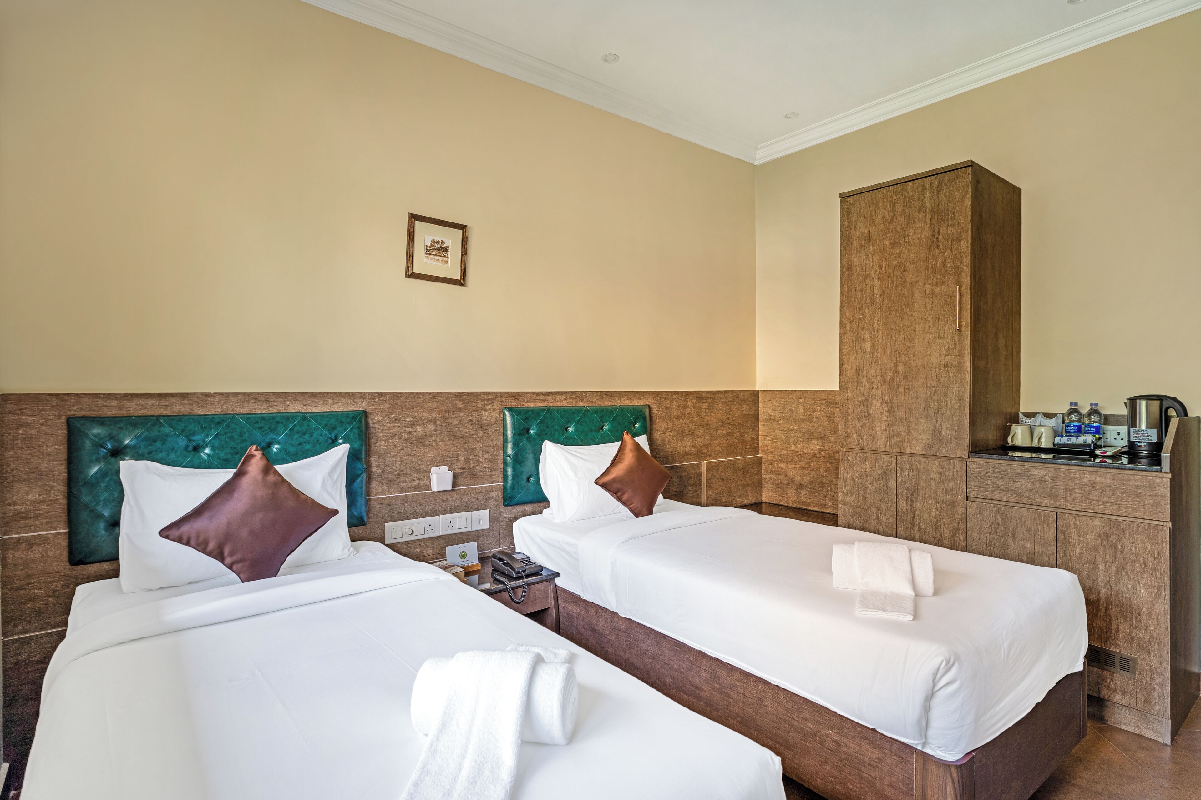 Habitación de hotel con dos camas, cada una con cabecera, ropa de cama blanca y almohada marrón. Hay un mueble de madera y una mesita con un teléfono y una botella.