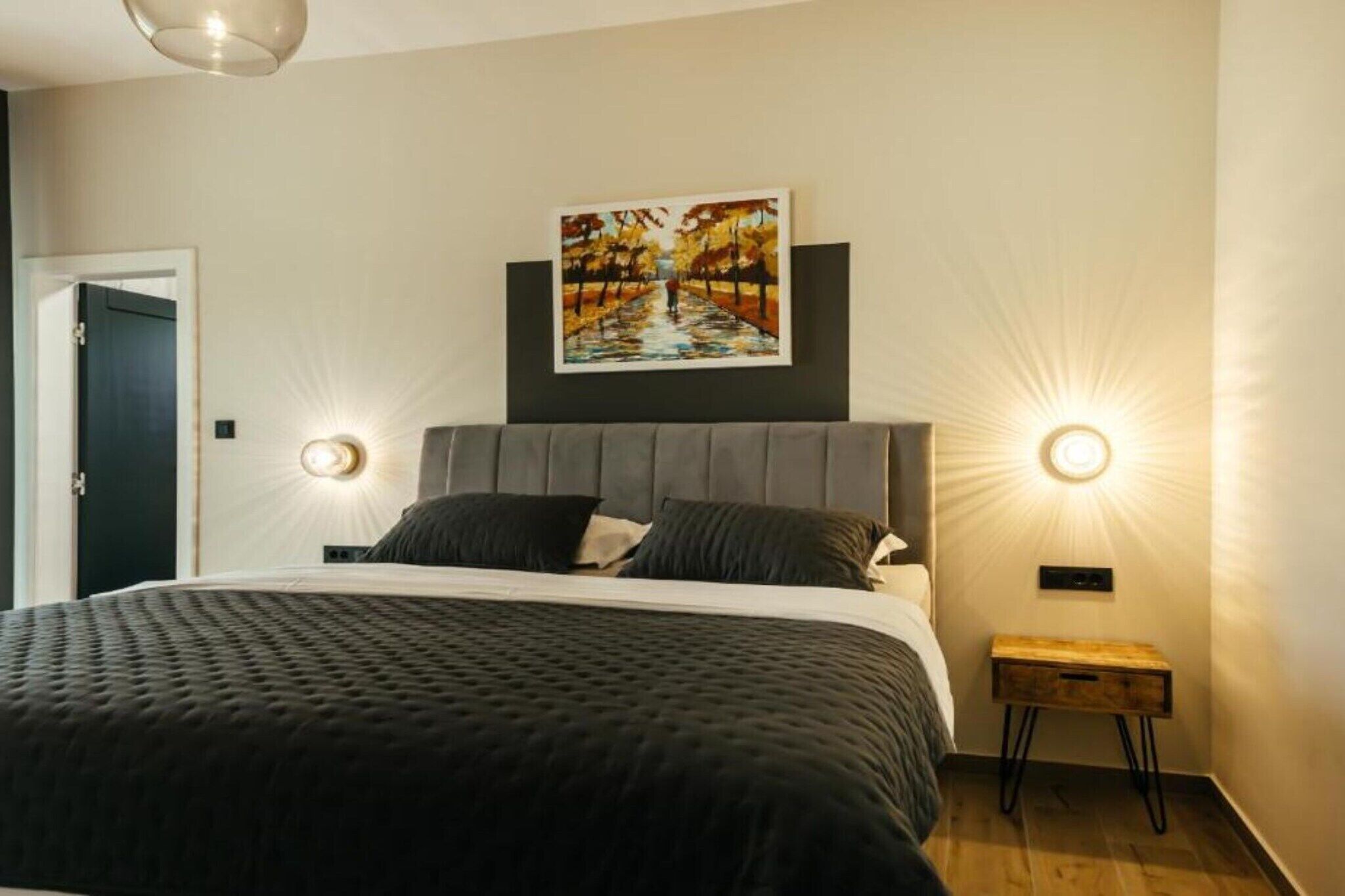 Ein modernes Schlafzimmer mit einem großen Bett, einer Kopfteil, zwei Wandleuchten, einem Gemälde über dem Bett und einem kleinen Nachttisch.
