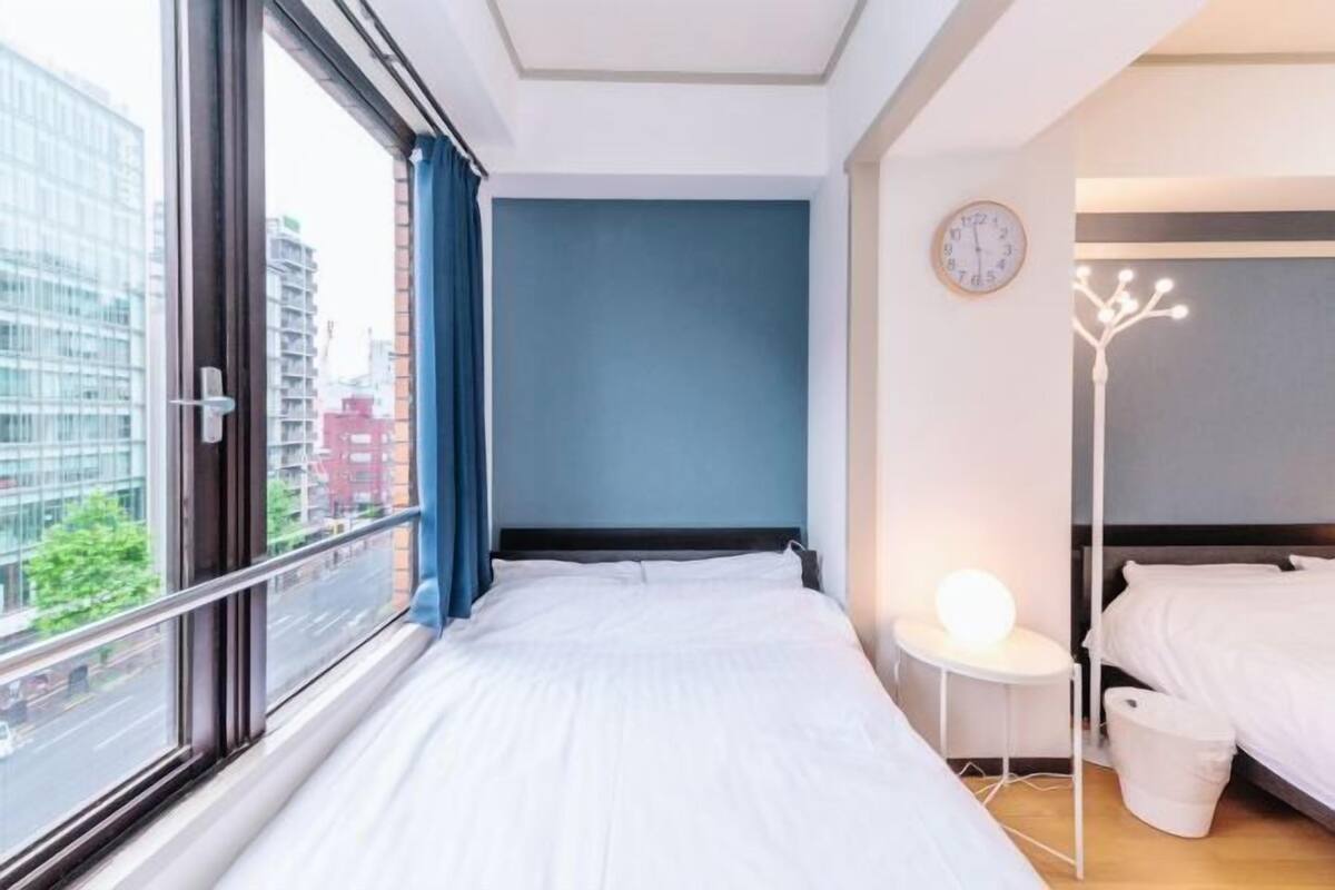 Habitación de hotel con un ventanal grande, una cama, una mesita de noche con lámpara y un reloj de pared.