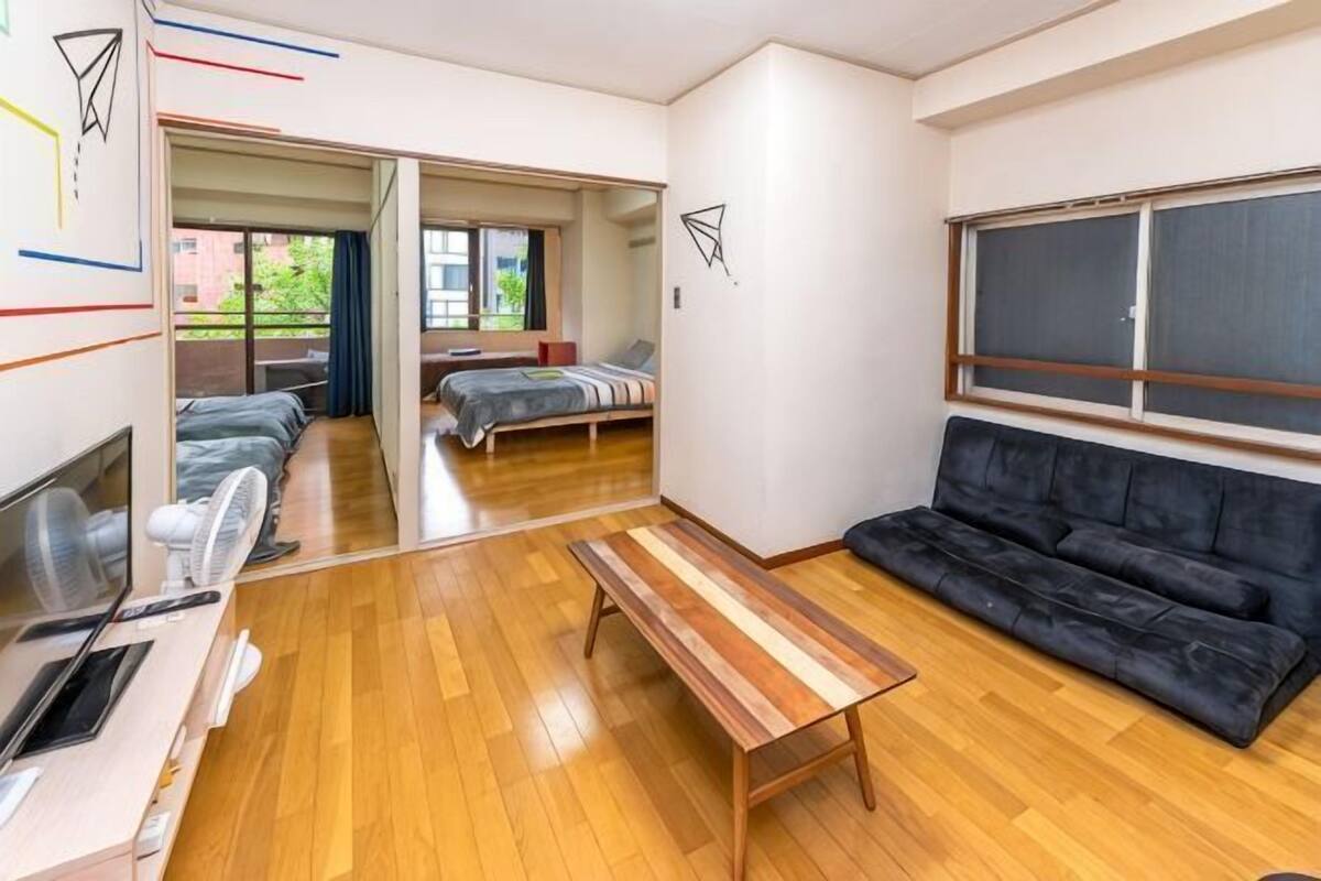 Una habitación con suelo de madera, un sofá negro, un banco de madera, un escritorio con una silla y una cama al fondo.