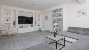 A modern living room with a grey sofa, a wooden chair, a coffee table with flowers, a flat-screen TV, and a decorative wall panel. - Countryside Villa Tajo - Four Bedroom Holiday Home  with Swimming Pool (Imotski)