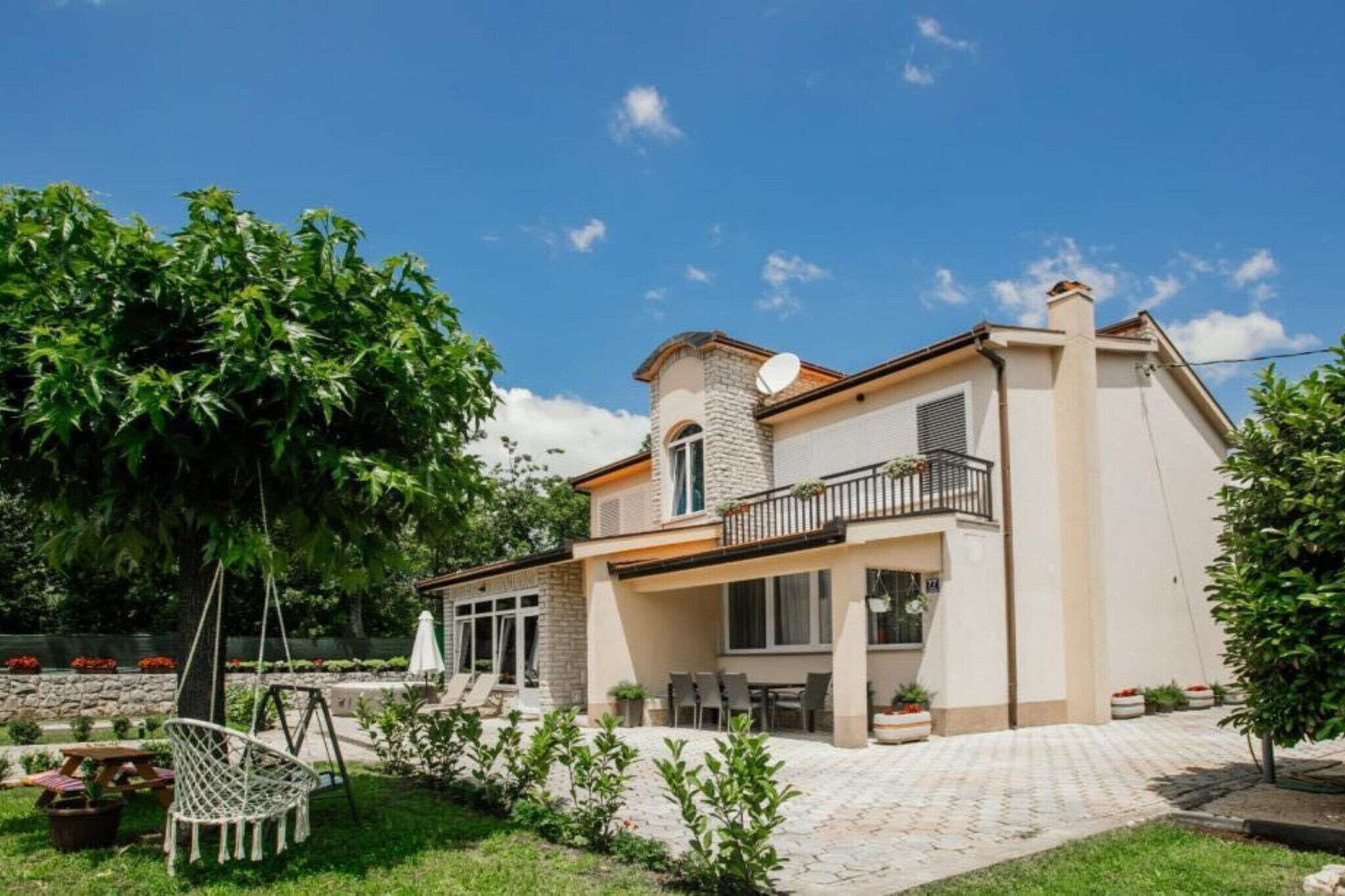 A two-story house with a stone facade, a balcony, and a garden with a swing chair.