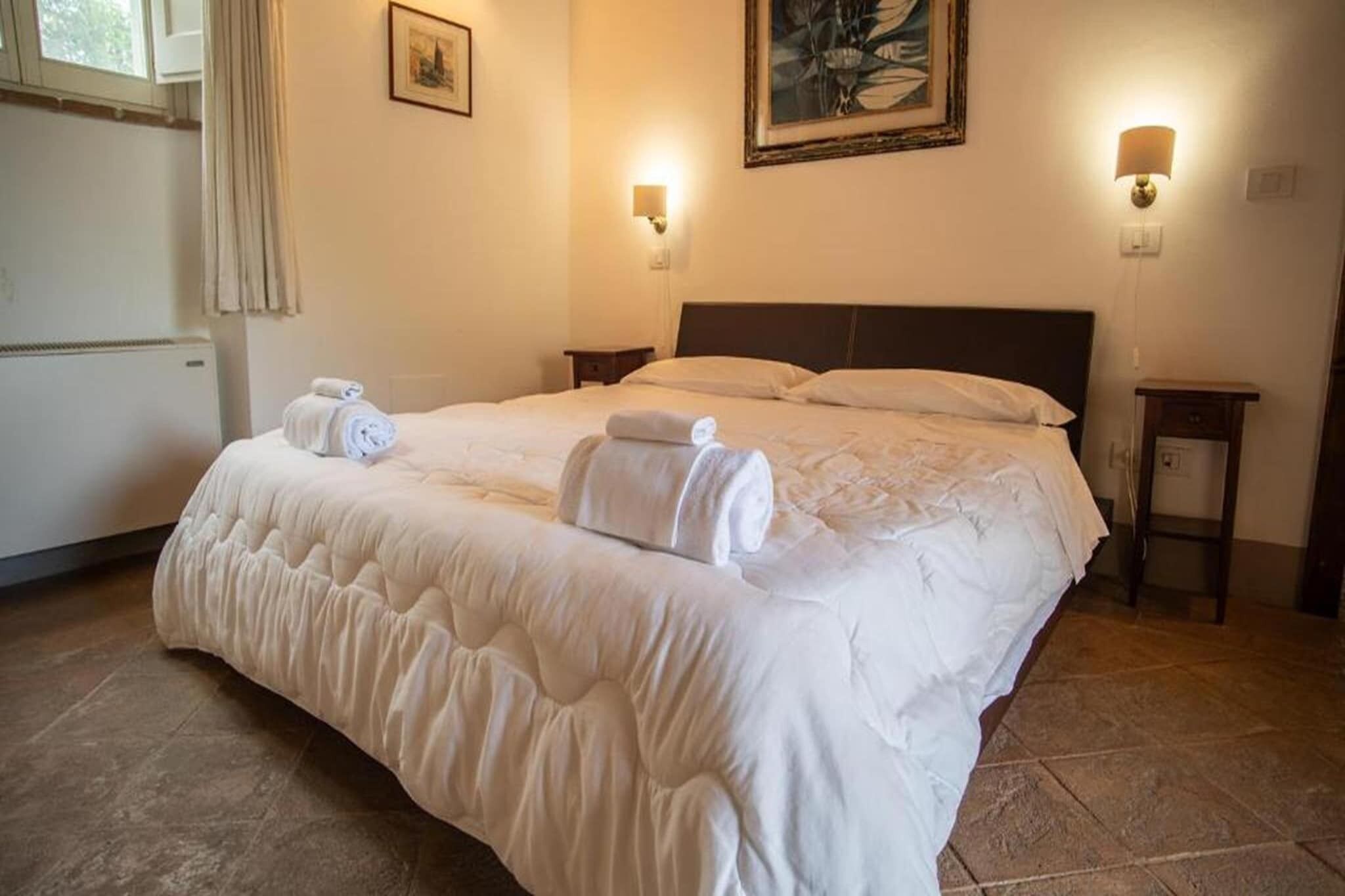 Een net opgemaakt bed met witte lakens en opgerolde handdoeken, een hoofdbord, nachtkastjes en wandlampen.