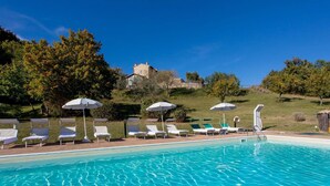 Une piscine avec des chaises longues et des parasols, un bâtiment en pierre en arrière-plan et un ciel bleu dégagé.