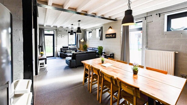 A modern kitchen with a wooden table and chairs, a black hanging light, and a view of the living area with a sofa and plants.