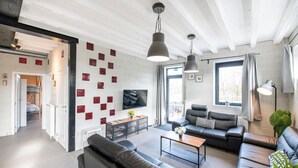 A modern living room with black leather sofas, a coffee table, and a TV on the wall. There are red square decorations on the wall, and a large window with curtains.