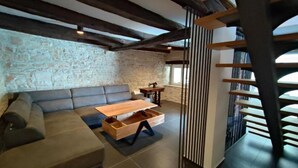 Una sala de estar moderna con vigas de madera a la vista, una pared de piedra, un sofá, una mesa de centro con cajones y una escalera con barandilla.
