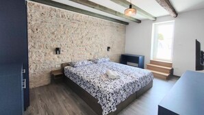 Un dormitorio con pared de piedra, cama con colcha floral, mesita de noche, una silla y ventana con vistas.