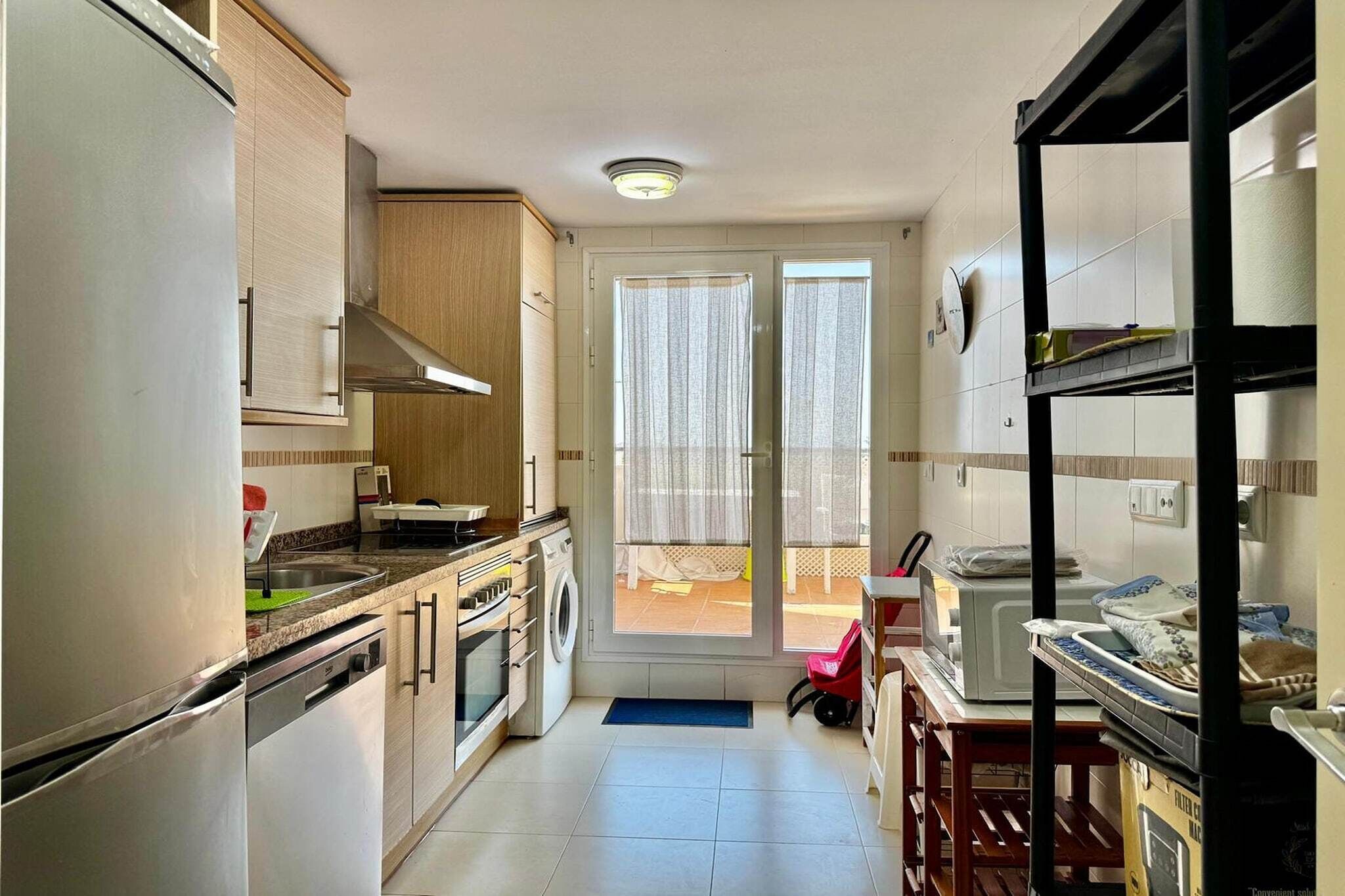 A compact kitchen with a refrigerator, oven, microwave, and washing machine. There is a window with curtains, a door leading outside, and a small table with a chair.