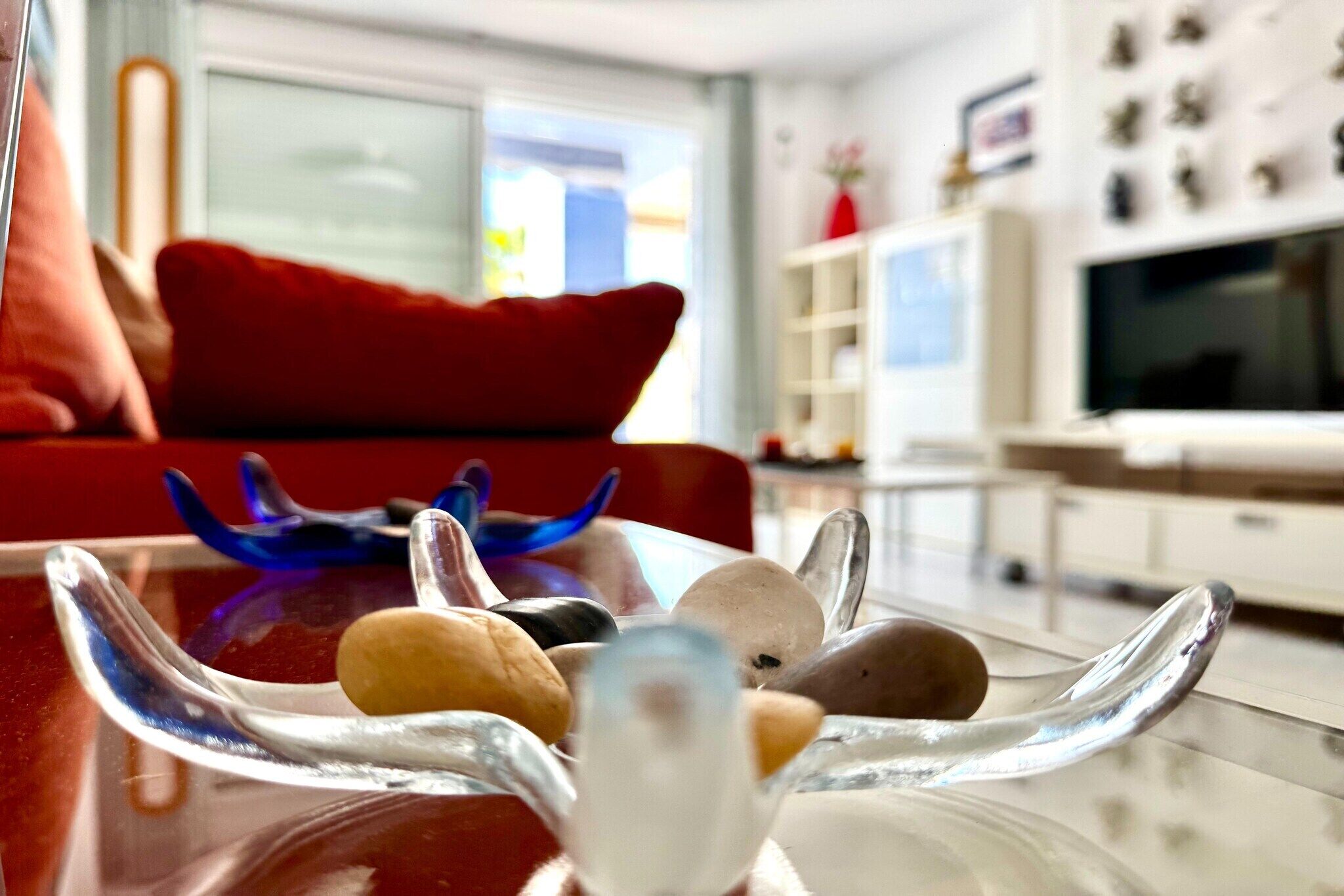 Ett modernt vardagsrum med ett soffbord i glas som pryds av dekorativa föremål, en röd soffa och en tv i bakgrunden.