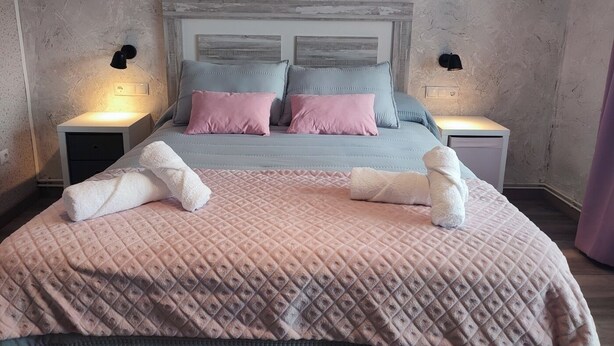 Ein ordentlich bezogenes Bett mit einer rosa Steude und zwei Kissen, ein weißer Nachttisch mit einer Lampe sowie ein ähnlicher Nachttisch auf der gegenüberliegenden Seite.