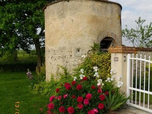 Une construction en pierre dotée d’une petite entrée en arc de cercle, entourée d’une grille métallique blanche et de plantes en fleurs.