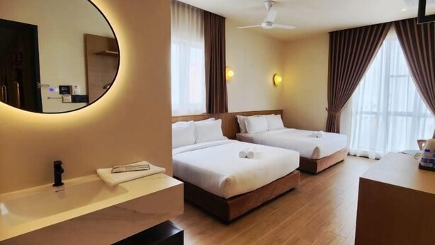 Habitación de hotel con dos camas, un espejo redondo, ventilador de techo, ventana grande con cortinas y mesita de noche con lámpara.