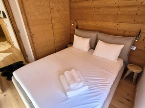 A neatly made bed with white linens and two pillows, a wooden headboard, and a small wooden stool beside the bed.