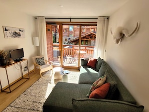 Un salon confortable avec un canapé, un téléviseur et une petite table. Il y a un balcon avec des meubles en bois et une vue sur les immeubles.