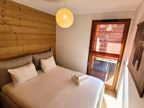 Une chambre à coucher avec une tête de lit en bois, un lit recouvert de draps blancs, un luminaire suspendu et une fenêtre donnant sur un immeuble.