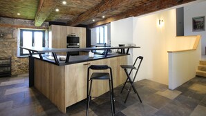 Une cuisine moderne avec un îlot central en bois, deux tabourets de bar, un four encastré et un mur en pierre.