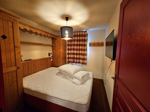 A bedroom with a bed, wooden paneling, a hanging light fixture, a plaid curtain, and a mounted television.