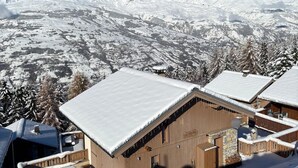 A wooden chalet with a snow-covered roof, set against a backdrop of snow-covered mountains and trees.