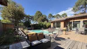 Eine Holzterrasse mit Gartenmöbeln, einem Pool und einem Gebäude mit großen Fenstern.