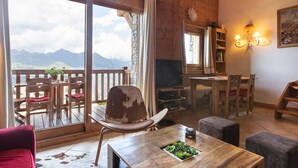 Un salon chaleureux avec un décor intérieur en bois, une grande fenêtre offrant une vue sur les montagnes, un coin repas avec des meubles en bois et un téléviseur posé sur un meuble en bois.