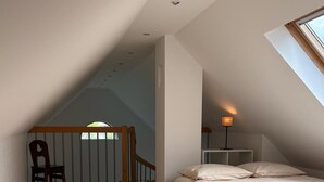 Een slaapkamer met een schuine kap, houten vloeren, een bed met witte beddengoed, een stoel en een nachtkastje met een lamp.