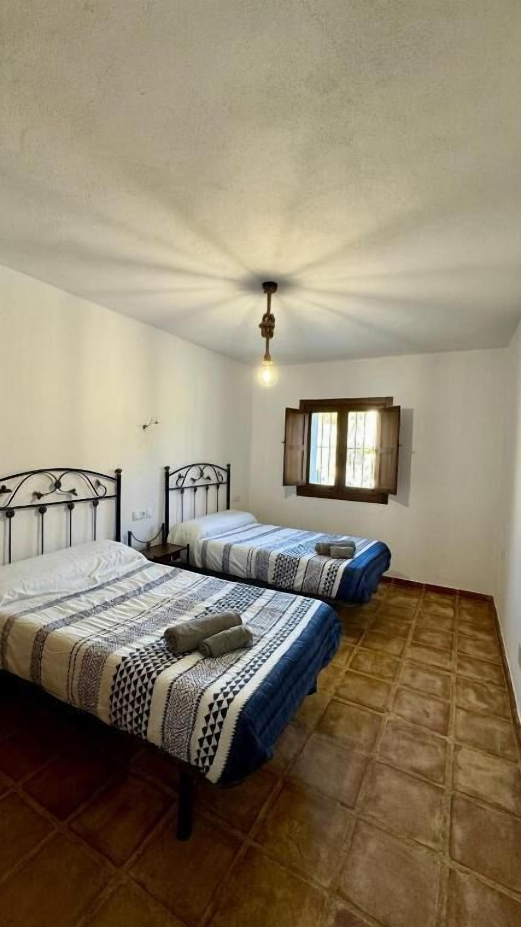 Una habitación con dos camas, ventilador de techo, ventana con cortinas y piso de baldosas.