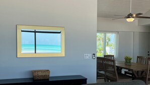 Un salon avec vue sur l’océan, visible par une fenêtre, doté d’un ventilateur de plafond et comprenant un coin repas avec une table et des chaises.
