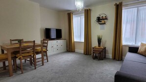 Una sala de estar con un sofá, un televisor sobre un mueble bajo, una mesa de comedor con sillas y una mesita auxiliar con una planta.