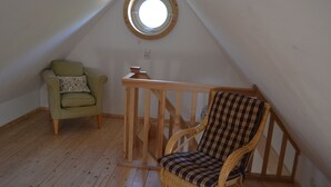 Huone, jossa on kalteva katto, puinen tuoli, rottinkituoli ja pyöreä ikkuna.