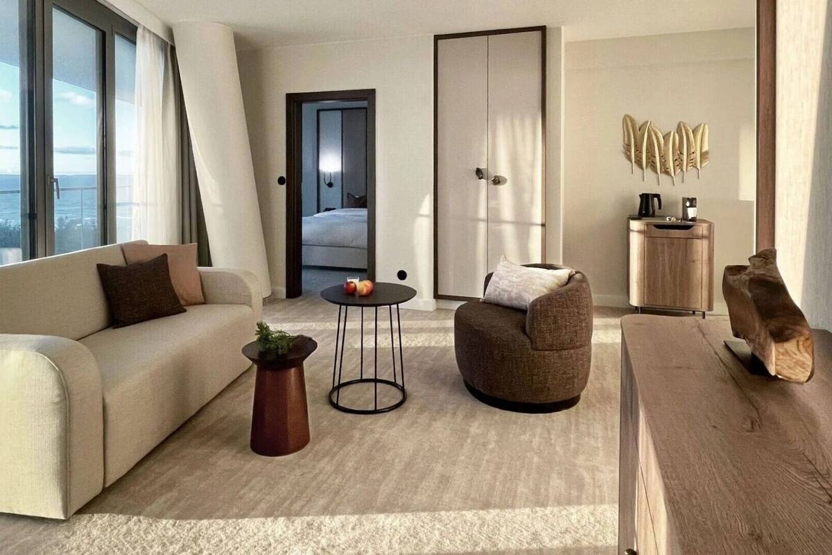 Ein modernes Wohnzimmer mit einer Couch, einem Sessel, einem Couchtisch und einem Holznachttisch. Durch eine Tür ist das Schlafzimmer zu sehen und es gibt ein großes Fenster mit Vorhängen.