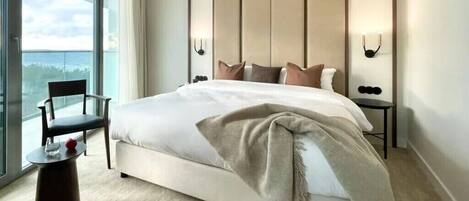 A modern hotel room with a large bed, a chair, a small table, and a view of the outdoors.