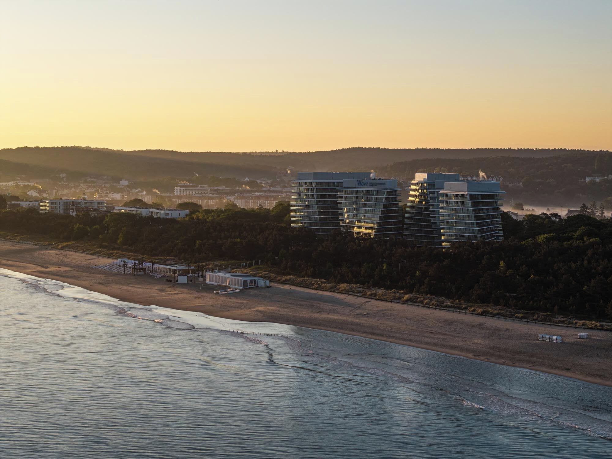 A coastal hotel with multiple high-rise buildings, a beach in the foreground, and a hilly landscape in the background.
