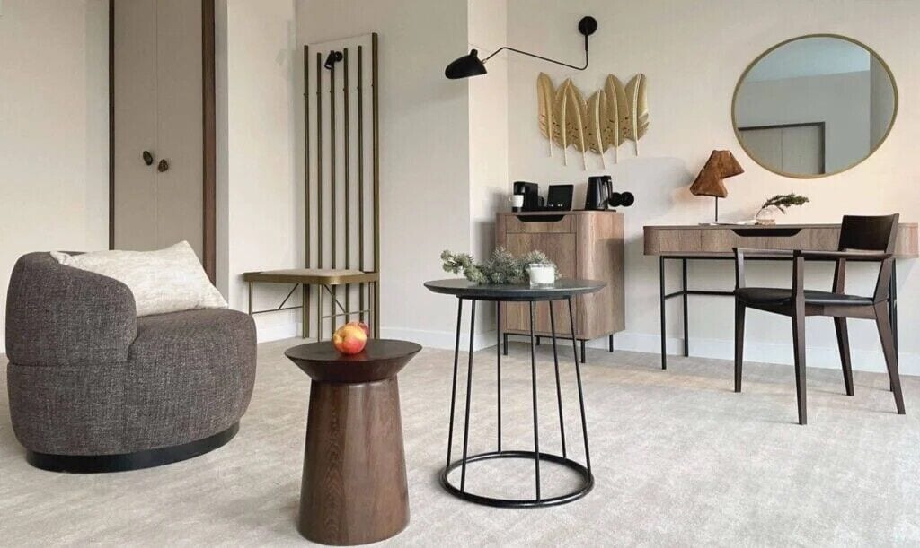Ein modernes Wohnzimmer mit einem grauen Sessel, einem runden Holztisch mit einem Apfel, einem kleinen Tisch mit einer Lampe, einem Schreibtisch mit Stuhl und einem großen runden Spiegel an der Wand.