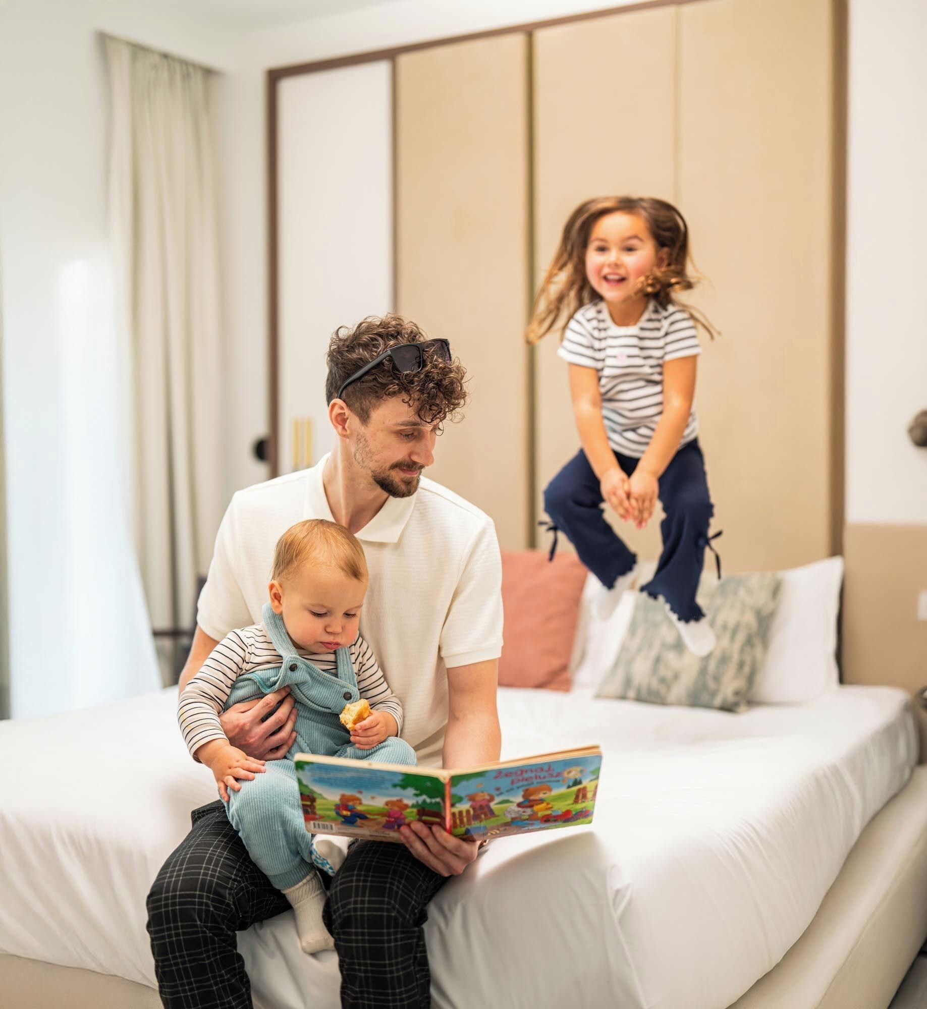 Ein Mann liest ein Buch vor für ein Baby, während ein Kind im Hintergrund spielt.