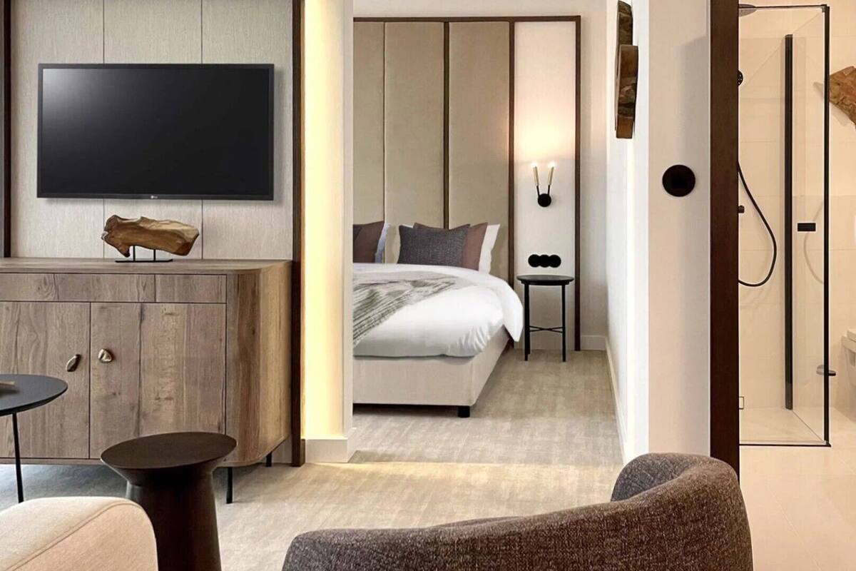 Ein modernes Hotelzimmer mit großem Flachbildfernseher, einem Holzschrank, einem runden Tisch, einer Couch, einem Bett mit Kissen und einer Duschkabine.