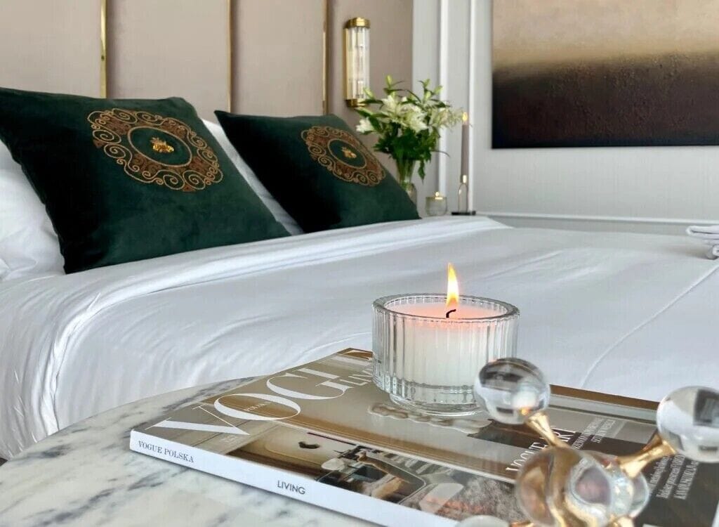 A neatly made bed with two pillows, a magazine, and a lit candle on a marble table.