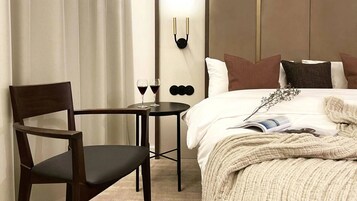 Habitación de hotel con cama, una silla, una mesita con dos copas de vino y una lámpara fijada a la pared.