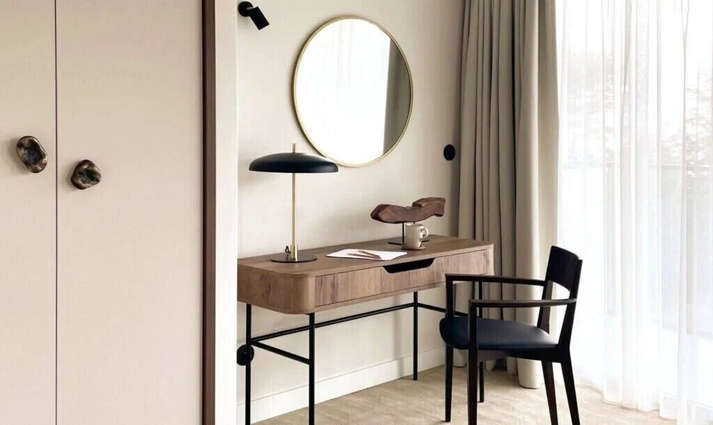 A minimalist room with a wooden desk, a chair, a round mirror, and a lamp.
