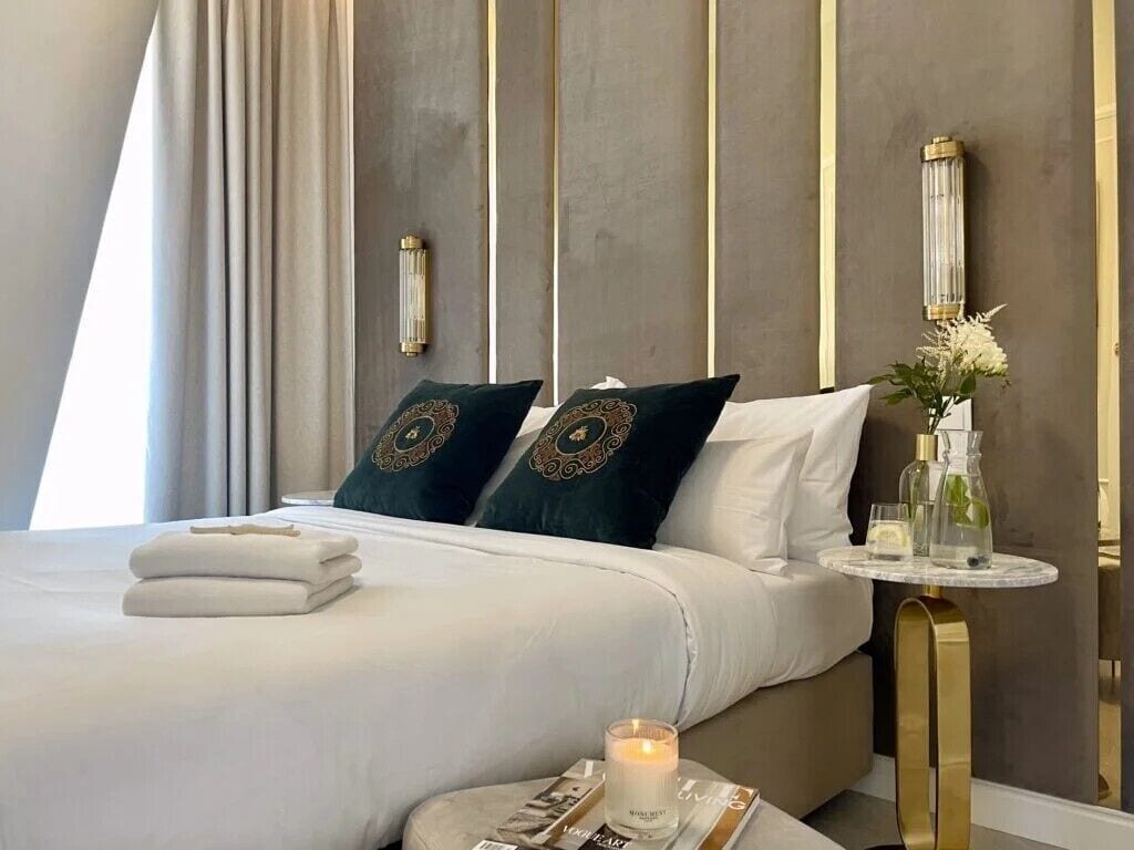 A neatly made bed with white linens and decorative pillows, a bedside table with a vase of flowers, and a magazine.