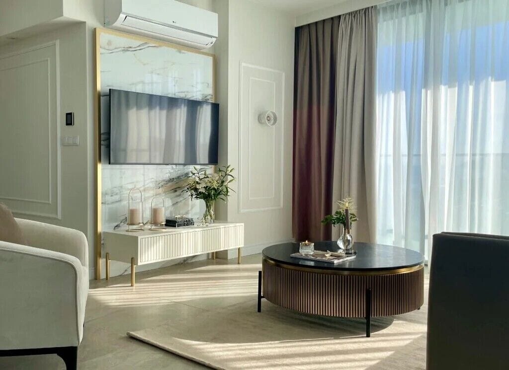 A modern living room with a round coffee table, a sofa, a TV mounted on a marble wall, and a large window with curtains.