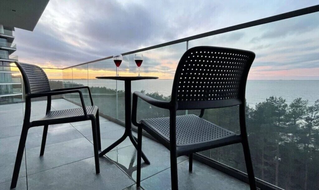 Un balcón con dos sillas y una mesa, dos copas de vino tinto y vistas al mar y a los árboles.
