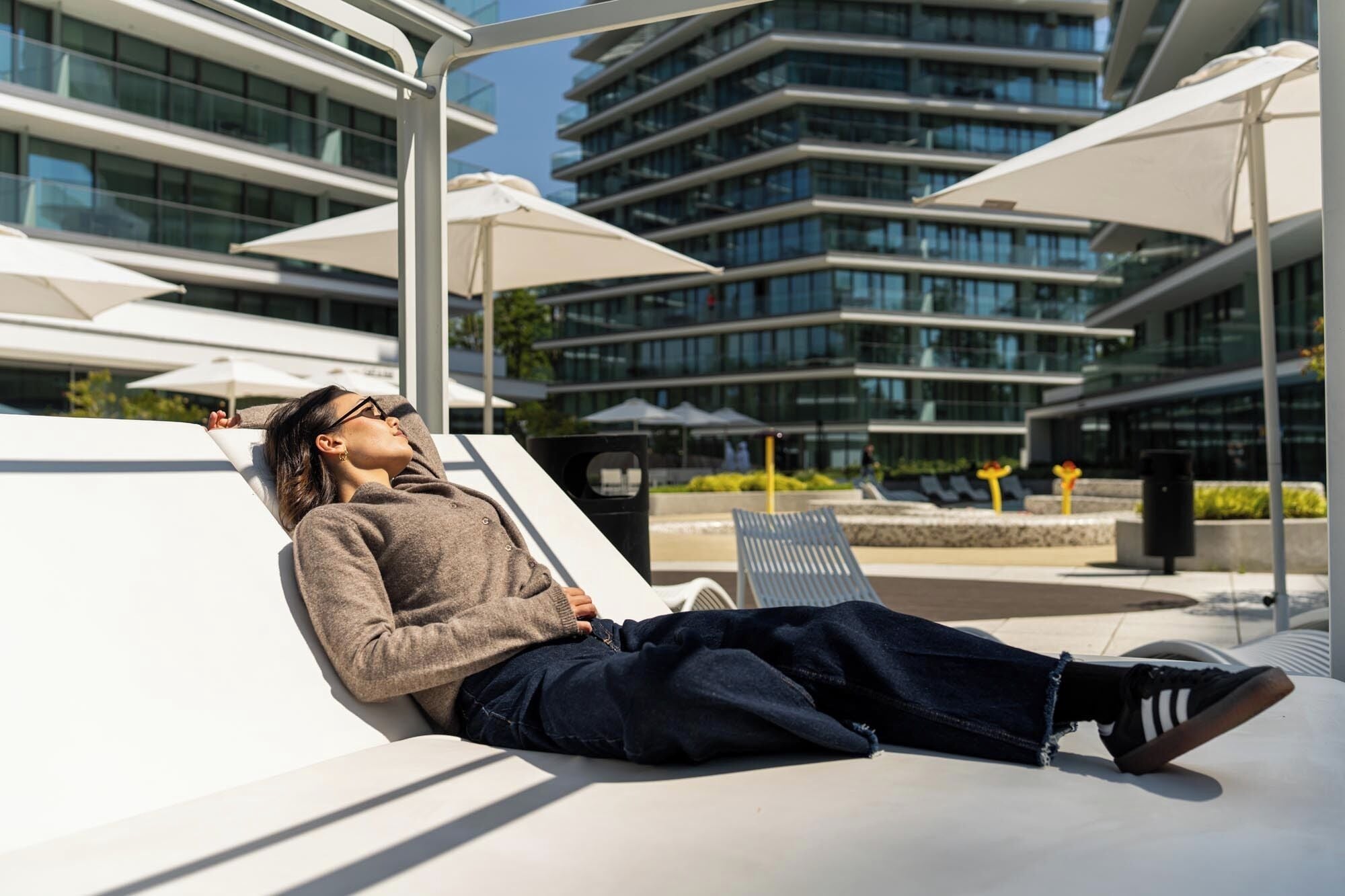 A person relaxing on a white lounge chair in an outdoor area with modern buildings in the background.