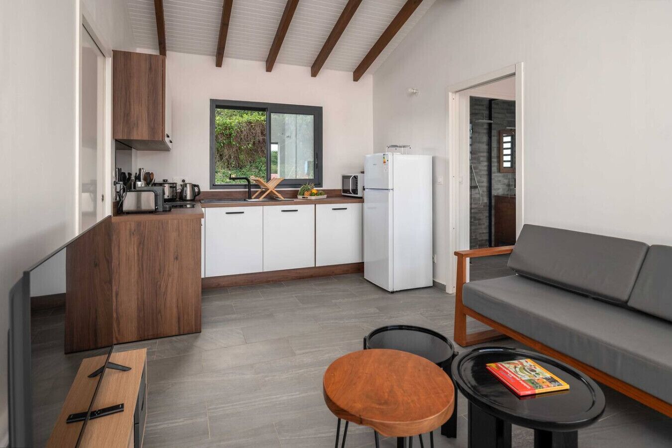 A compact kitchen with a refrigerator, microwave, and wooden cabinets. There is a dining area with a gray sofa and a small round table.