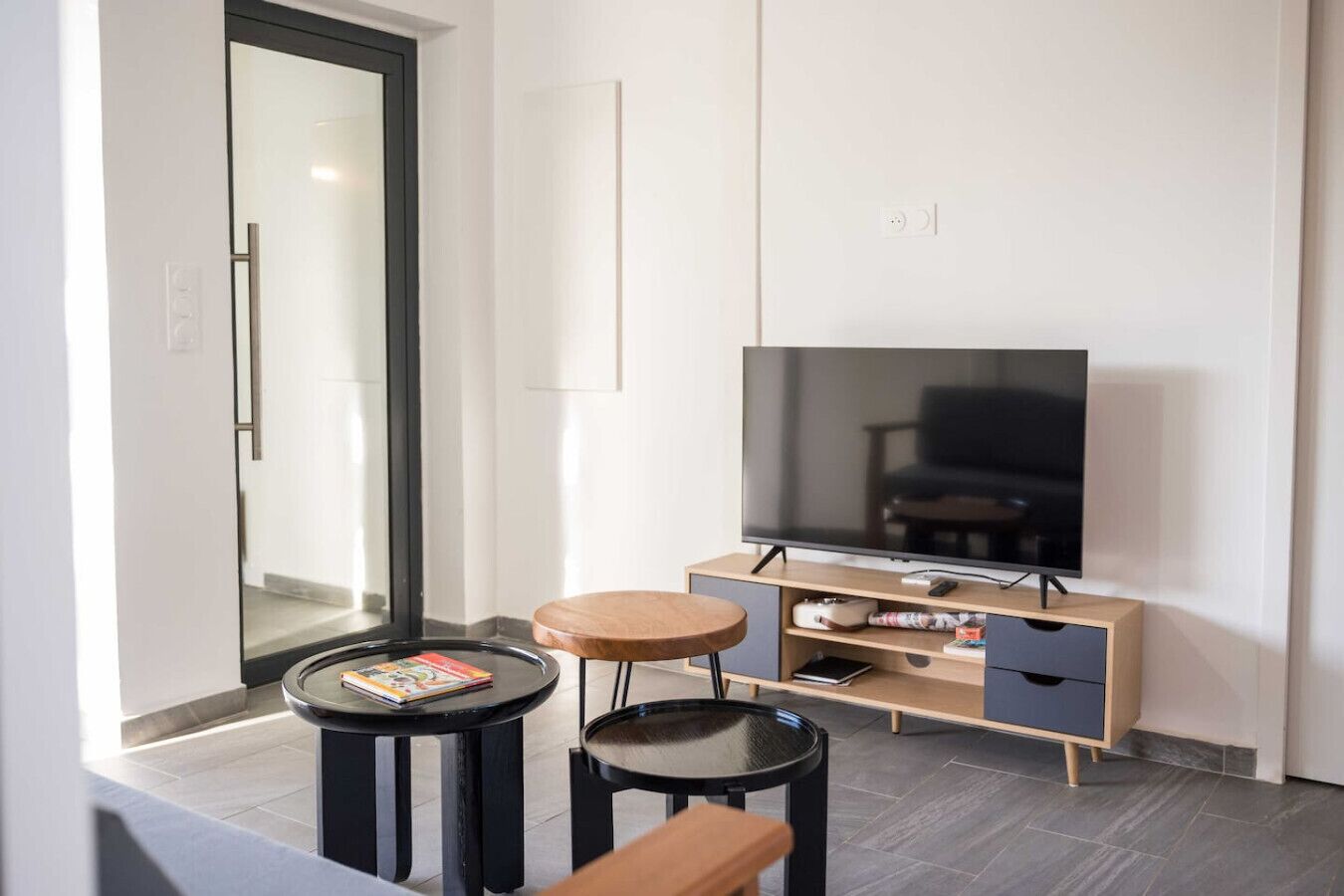 Een moderne woonkamer met een grote flatscreen televisie, een houten kast met lades en twee kleine ronde tafels met boeken.