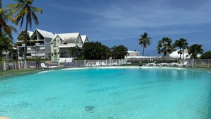 Une piscine d’un bleu éclatant, entourée d’une clôture blanche et de chaises longues, bordée de palmiers et d’un bâtiment doté d’un toit blanc.
